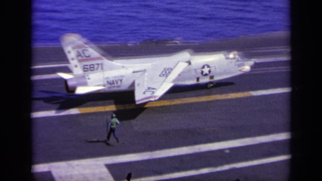 Navy jet sits on the carrier deck while a crew member walks near the aircraft.