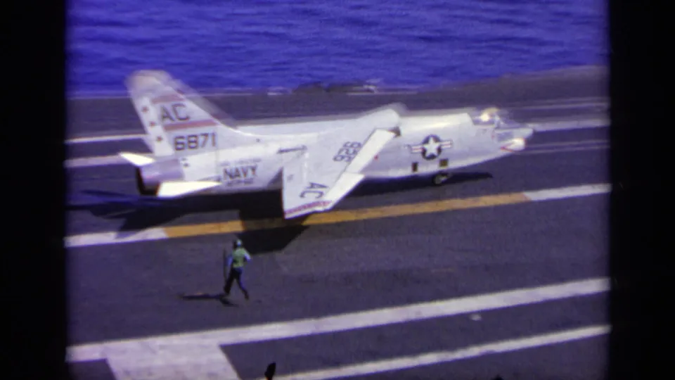 1963 Greece carrier-deck frame with a marked U.S. Navy jet and a green-clad deck crew member moving nearby.