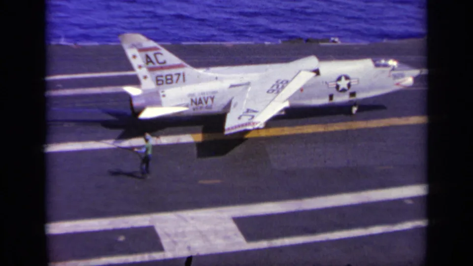 Home-movie frame showing a green-clad crew member near a white U.S. Navy jet on an aircraft carrier deck in 1963 Greece footage.
