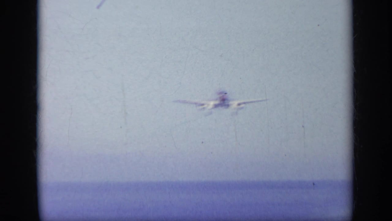 1963 Greece metadata frame showing a distant naval jet approaching over the sea in soft 8mm color.
