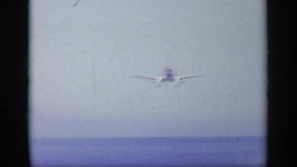 1963 Greece metadata frame showing a distant naval jet approaching over the sea in soft 8mm color.