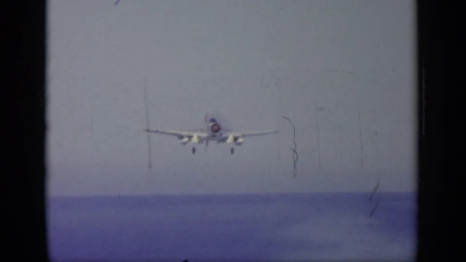 Head-on view of a naval jet approaching over open ocean, with landing gear lowered and 8mm film wear visible.