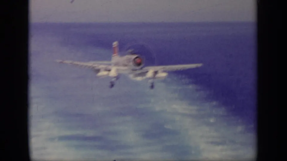 Closer 8mm frame of a naval jet on final approach over the sea, with wake-like water texture behind it.