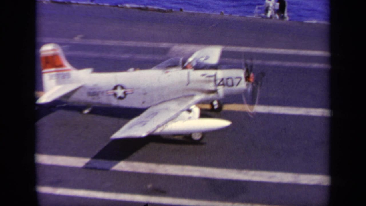 White U.S. Navy propeller aircraft taxiing on an aircraft carrier deck.