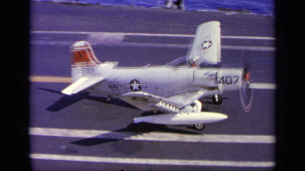U.S. Navy aircraft on carrier deck with one wing raised upward.