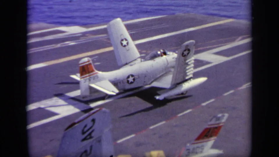 The white U.S. Navy aircraft continues across the marked carrier deck, with folded wings, ocean beyond, and another tail partially visible.