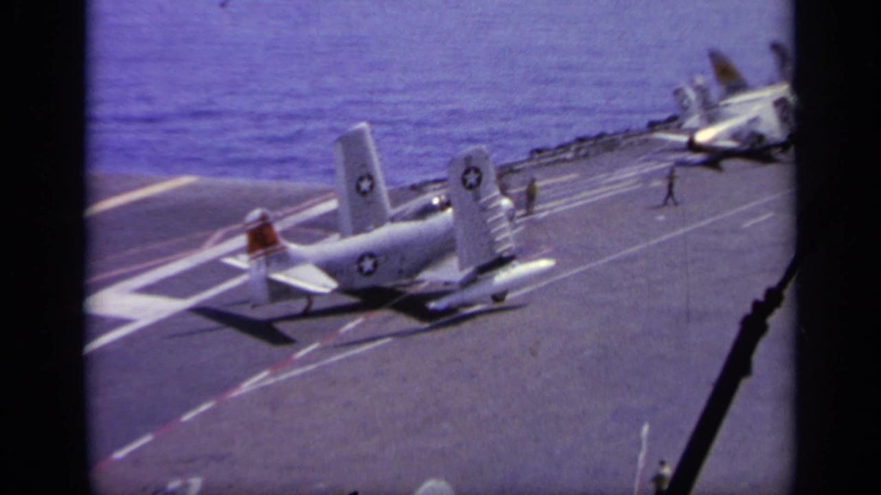 Navy aircraft on carrier deck with parked planes and crew in the distance.