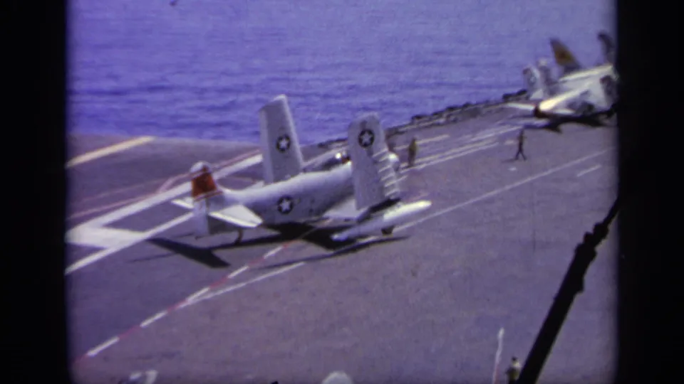 A white U.S. Navy aircraft taxis on the flight deck while other planes and crew are visible farther forward.