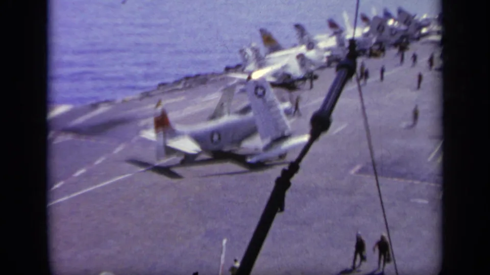 From an elevated viewpoint, the footage shows a Navy aircraft on deck with crew and several parked aircraft nearby.