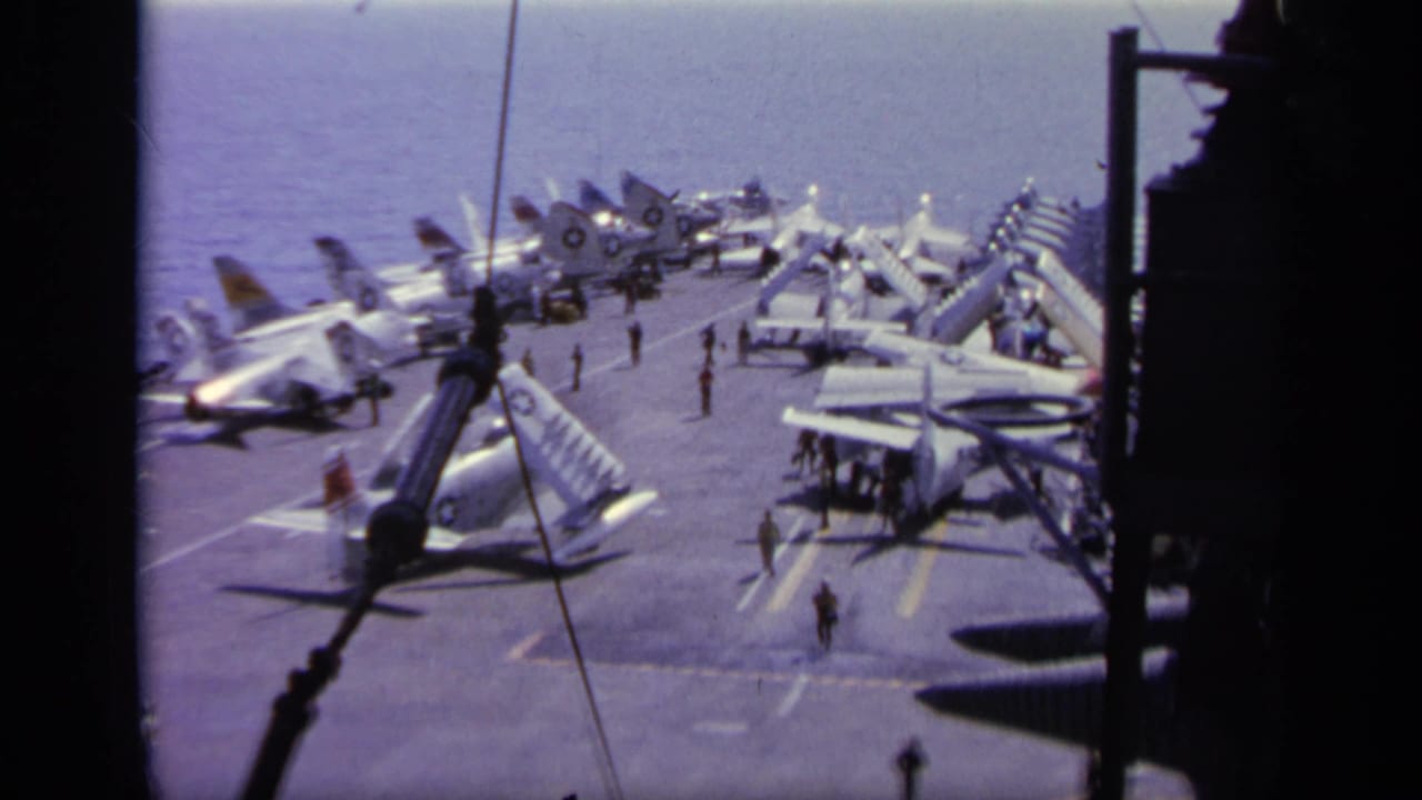Carrier flight deck with multiple parked jets and crew figures near the bow.