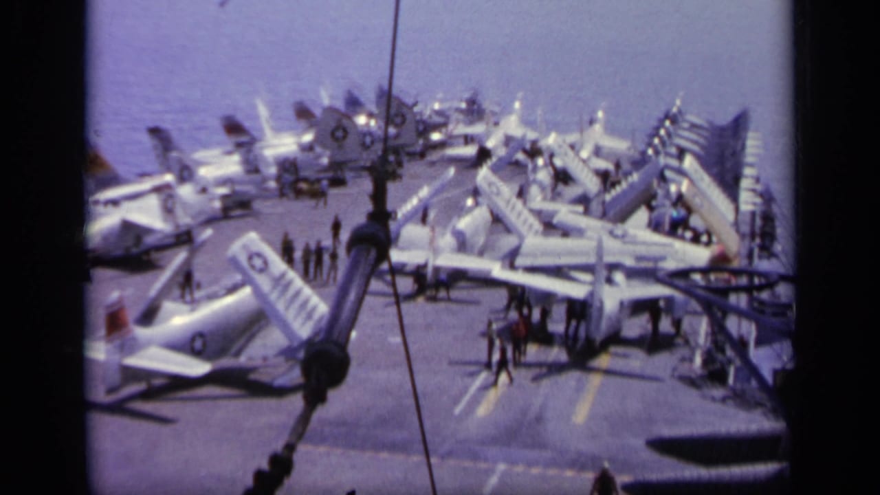 Aircraft carrier deck crowded with parked jets and small crew figures at sea.