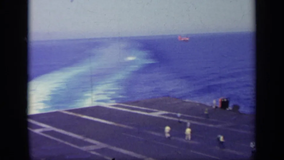 Handheld 8mm view across a carrier deck toward the sea, with tiny deck figures and a distant vessel-like shape.