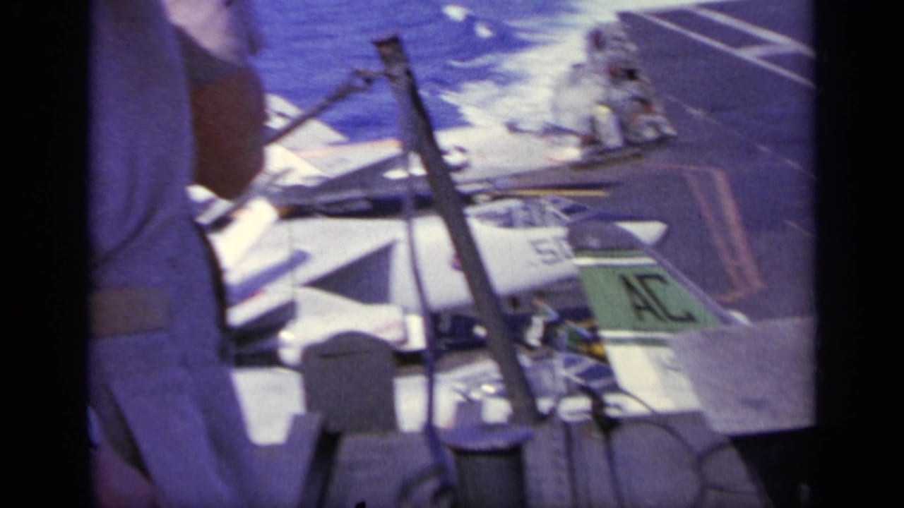 Aircraft carrier deck with parked Navy jets and a crew member partly visible at left.