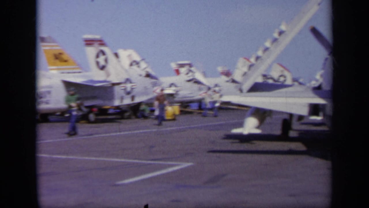 1963 8mm frame from Greece showing a line of U.S. Navy jets on a carrier deck under bright sun, with crew nearby.