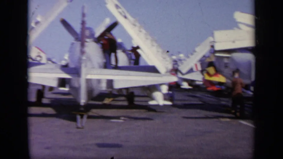 1963 8mm frame from Greece showing the rear of a Navy aircraft with folded wings, nearby stairs, and crew on the carrier deck.