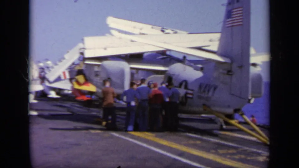 1963 8mm frame from Greece showing crew gathered beside a U.S. Navy aircraft on a carrier deck, with flag markings visible.