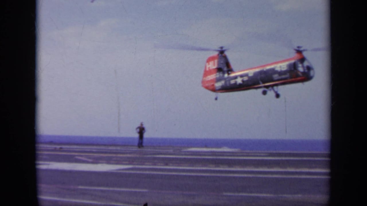 Red and blue tandem-rotor helicopter hovering above an aircraft carrier deck.