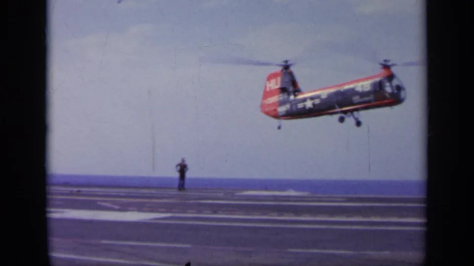 A red and blue U.S. Navy-marked tandem-rotor helicopter hovers over the carrier deck during the 1963 Greece footage.