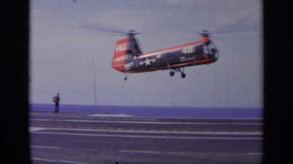 1963 Greece carrier footage shows the marked helicopter descending toward the flight deck as a deck figure watches from a distance.