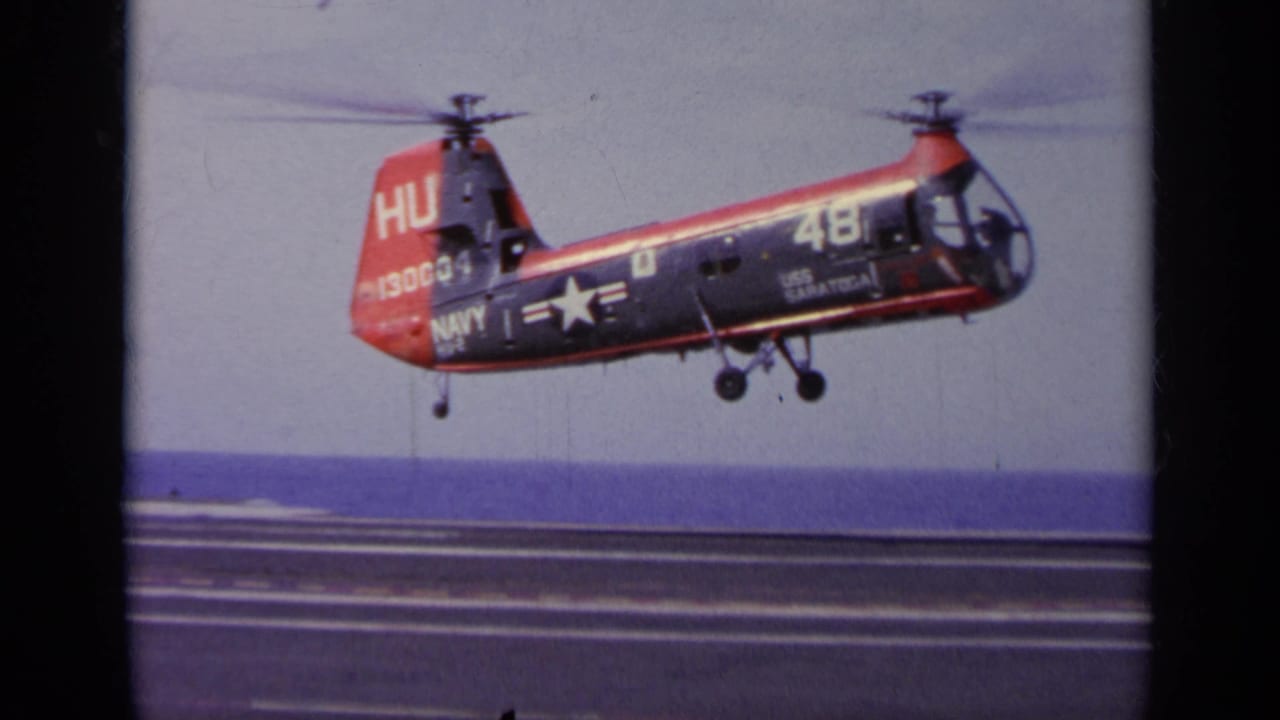 Red-and-blue U.S. Navy aircraft hovering above a carrier deck with ocean behind it.