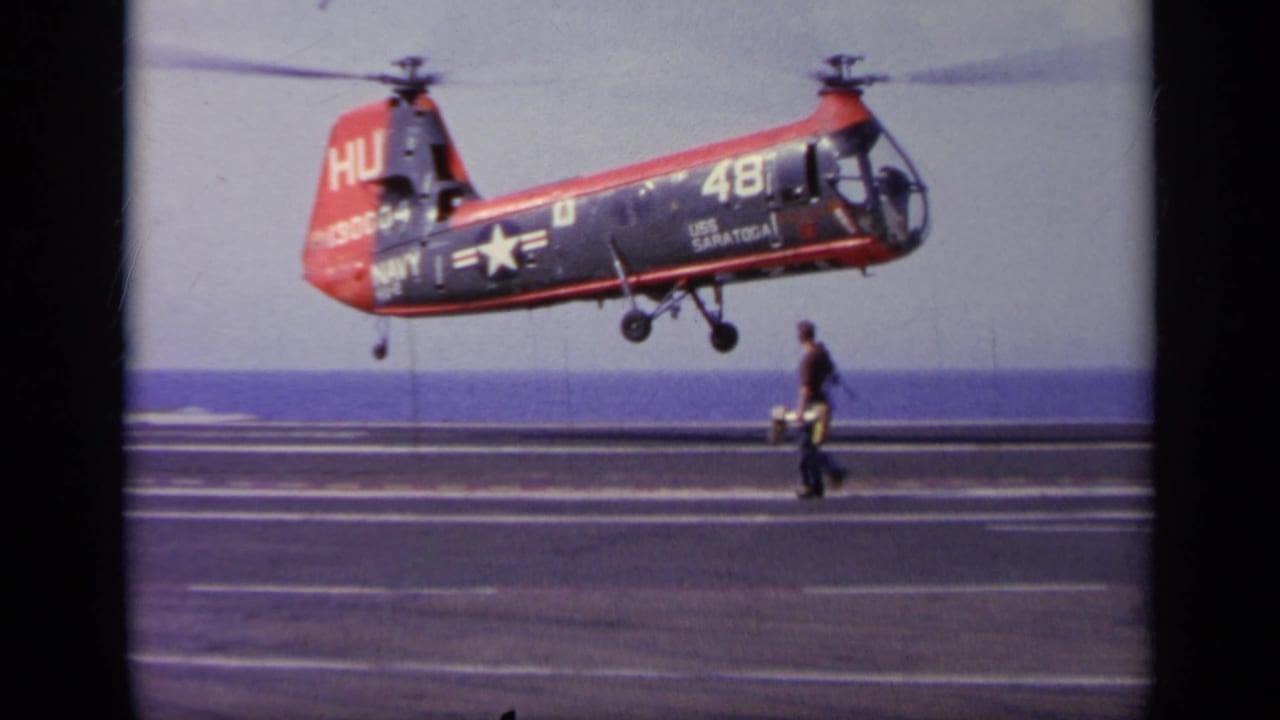 Navy aircraft hovering above the carrier deck as a deck crew member walks nearby.