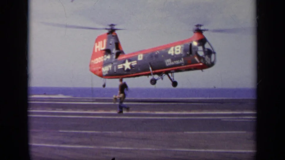 The U.S. Navy aircraft descends close to the carrier deck, with landing gear visible and a crew member moving across the foreground.