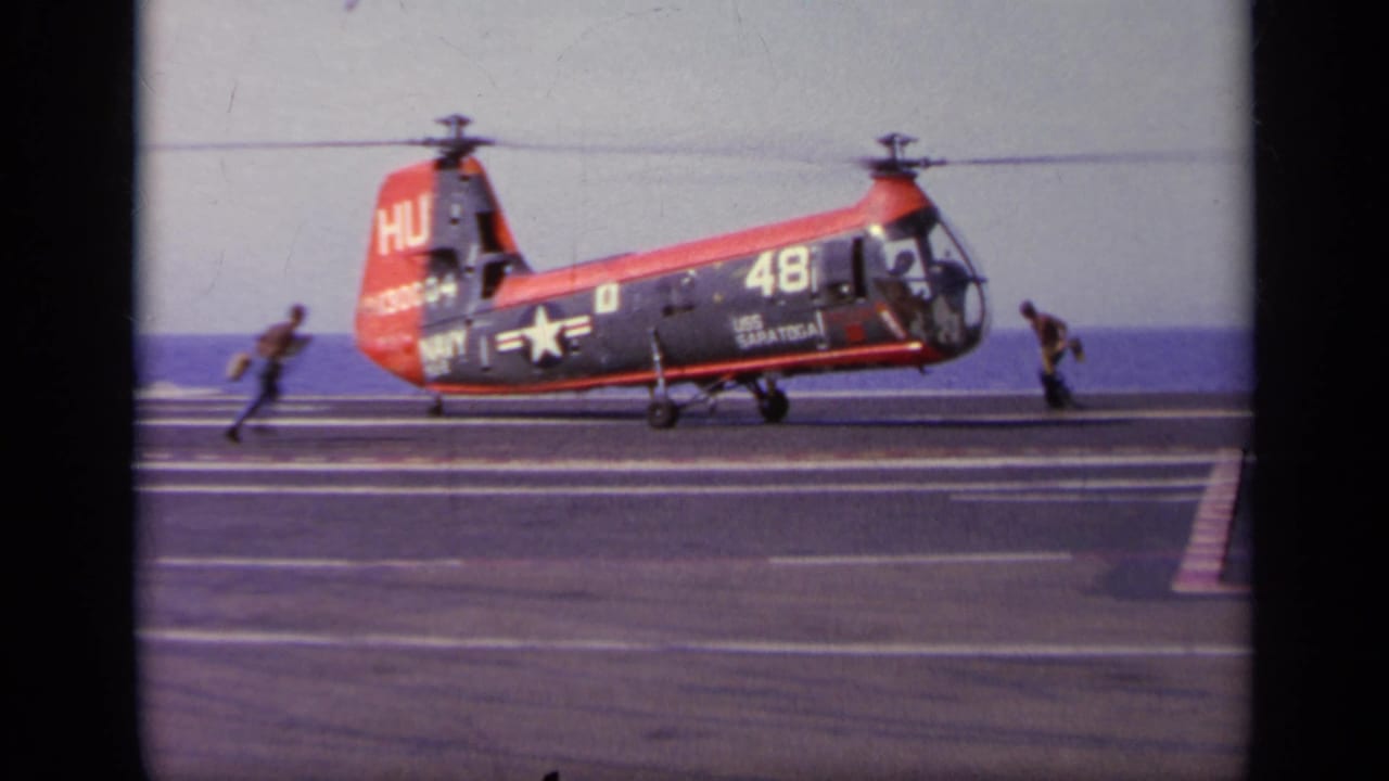 Red and dark Navy rotorcraft on an aircraft carrier deck with crew nearby and ocean behind it.