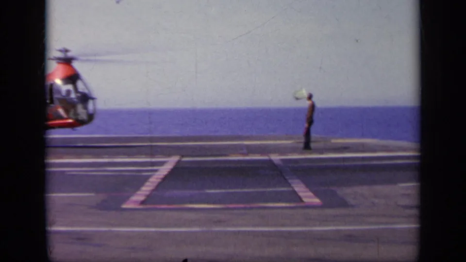 Carrier-deck crewman appears to signal near a partially visible Navy rotorcraft, with the ocean horizon behind him.