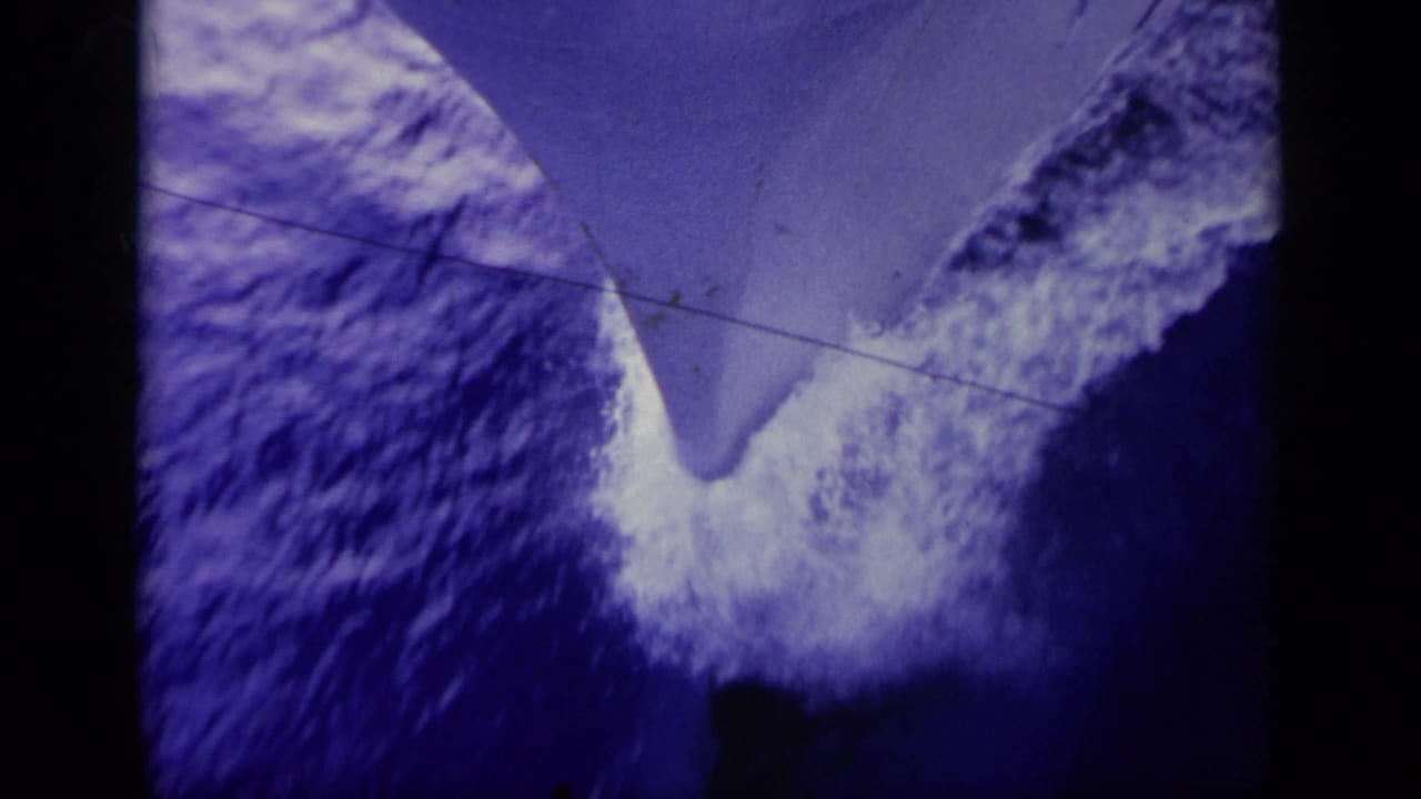 1963 Greece 8mm frame showing a boat bow or keel slicing through blue sea, with white foam and a horizontal scratch line.