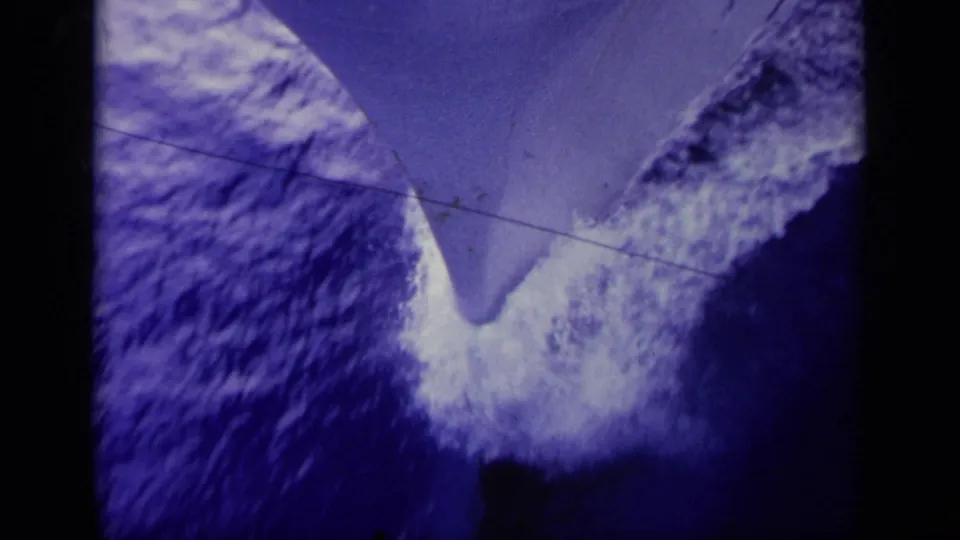 1963 Greece 8mm frame showing a boat bow or keel slicing through blue sea, with white foam and a horizontal scratch line.