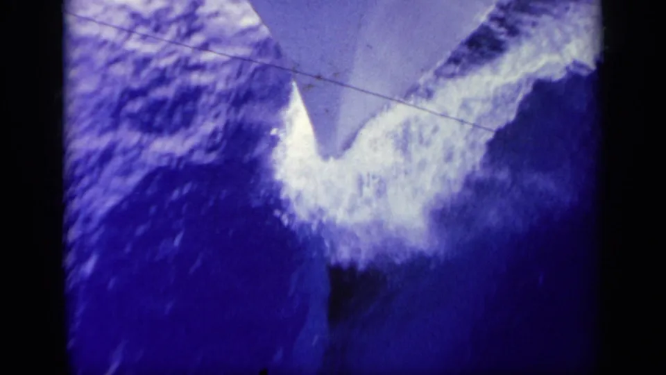 1963 Greece home-movie still of the boat prow area pushing through deep blue water, with white foam and analog grain.
