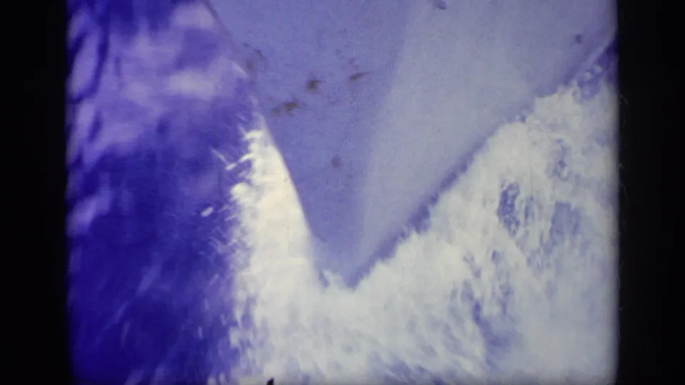 Close 8mm frame from Greece showing bright bow spray and a sharply angled boat edge over pale blue water.
