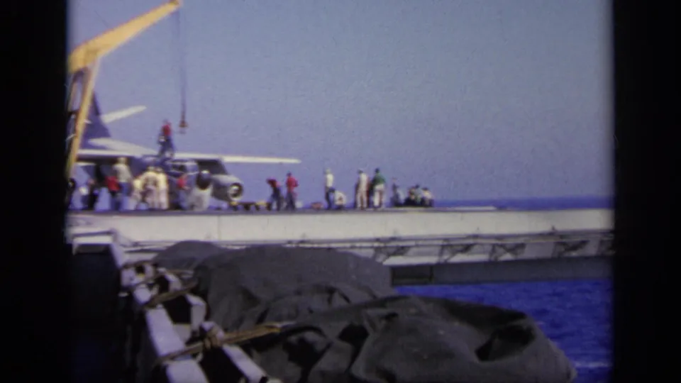 1963 Greece 8mm view of a carrier deck with crew gathered near a propeller aircraft and yellow crane beside the open sea.