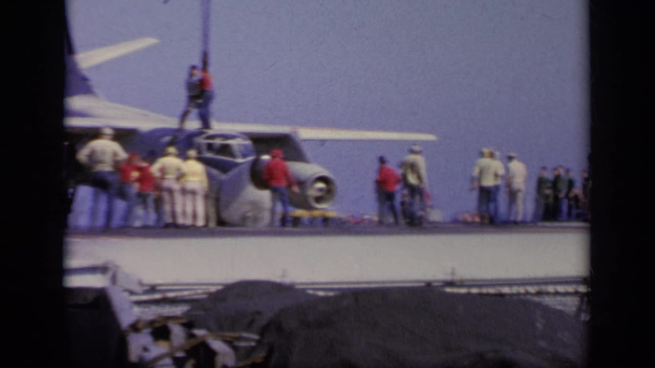 Gray military aircraft on a carrier deck with crew and observers nearby.
