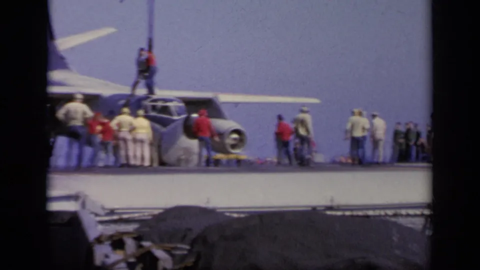 1963 Greece home-movie frame showing a gray military aircraft on a carrier deck as crew and observers stand around it.