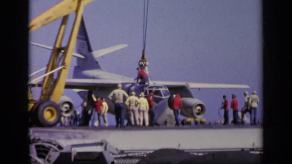 Yellow crane and hoist lines operate beside a gray military aircraft on a carrier deck in the 1963 Greece footage.