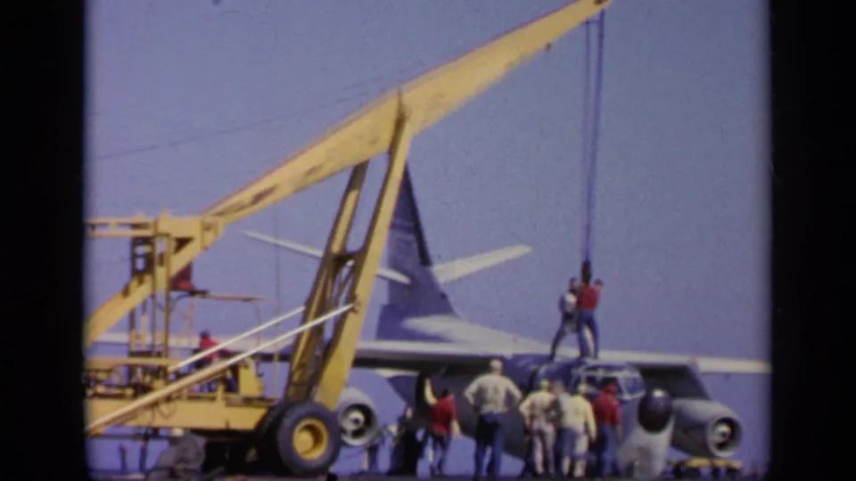 Carrier-deck crew watch as a crane boom and lines extend over the aircraft in this 1963 Greece home-movie frame.