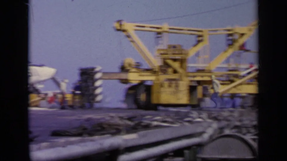 Wide home-movie view of a yellow deck crane, cables, equipment, and part of a military aircraft on a carrier deck in Greece.