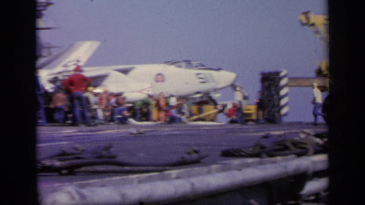 White Navy jet on an aircraft carrier deck with crew nearby.