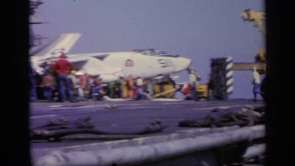 1963 8mm frame showing a white Navy jet on a carrier deck, with flight deck crew gathered near the nose and wing.