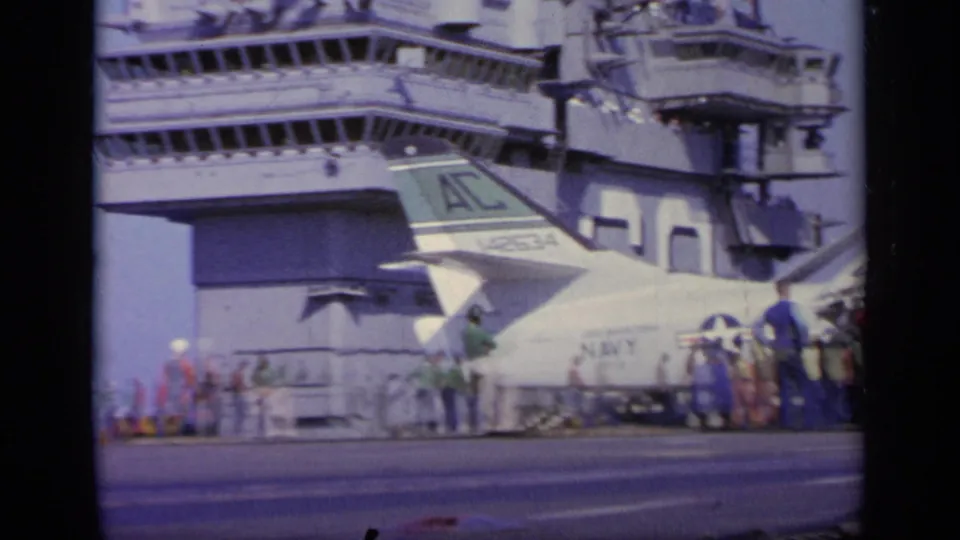 Carrier island and radar structure rise behind a white Navy aircraft tail, with deck crew visible along the flight deck.