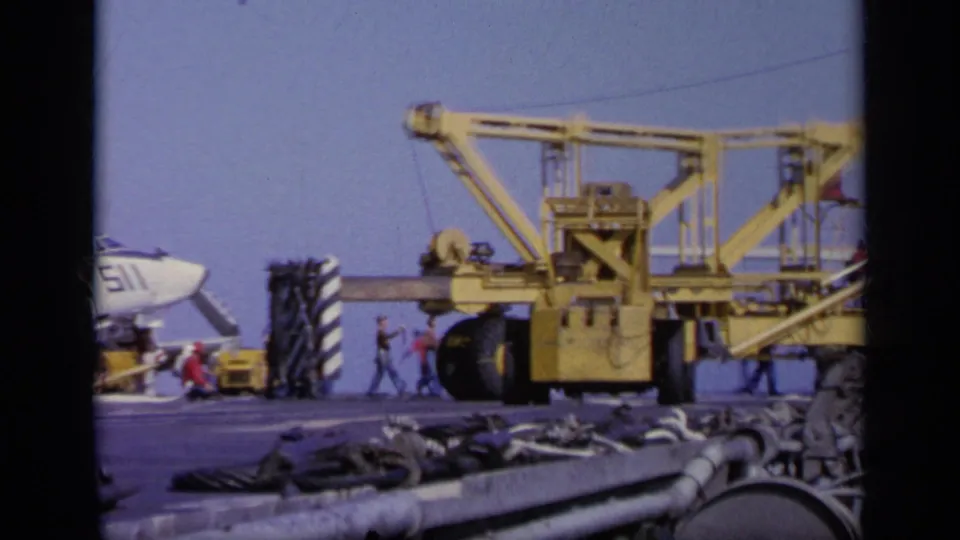 A yellow deck crane or support vehicle sits near a white jet on the carrier flight deck, with crew members moving nearby.