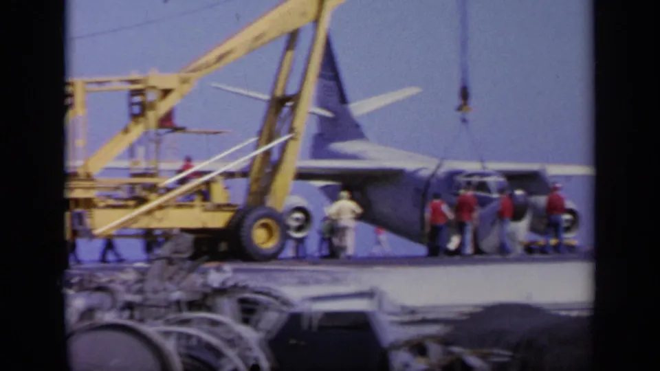 A yellow crane stands beside the rear section of a white military aircraft while deck crew work around the carrier deck.