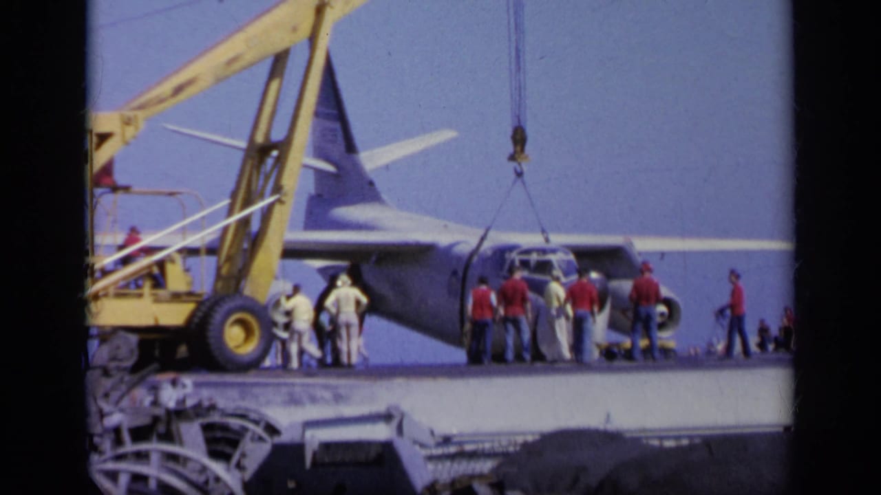 Transport plane on a ship deck near a yellow crane with crew gathered at the nose.
