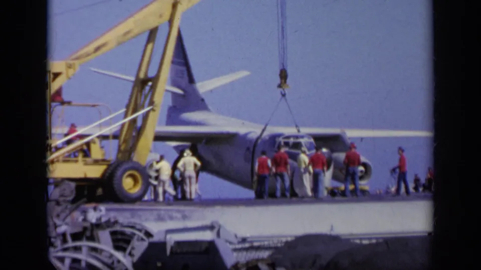 1963 Greece 8mm frame showing a military transport plane on a naval deck as workers stand near the nose and crane rigging.