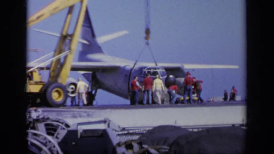 Distant 1963 Greece frame of a military transport aircraft on a warship deck with crew and crane equipment around it.