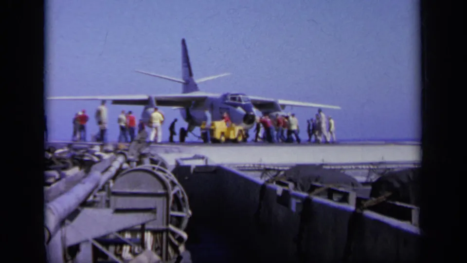 Wide 1963 Greece 8mm frame showing a military transport plane on a naval deck, surrounded by workers and foreground ship hardware.
