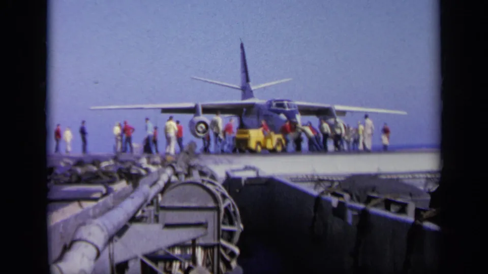 Military aircraft on a carrier deck in the supplied 1963 Greece footage, with people standing near the landing gear and tow tractor.