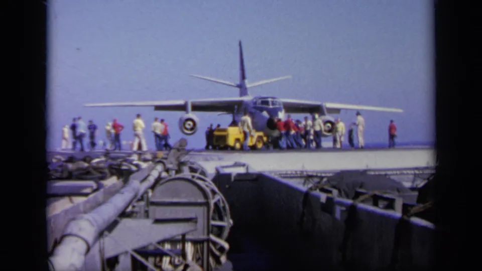 Distant 8mm view of a military aircraft on a carrier deck, with deck crew and visitors clustered near a yellow towing vehicle.
