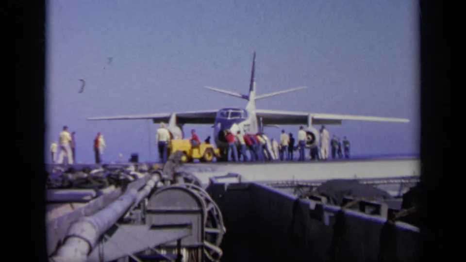 Front-facing carrier-deck view of the military aircraft, with personnel around the tow tractor and the ocean horizon behind it.